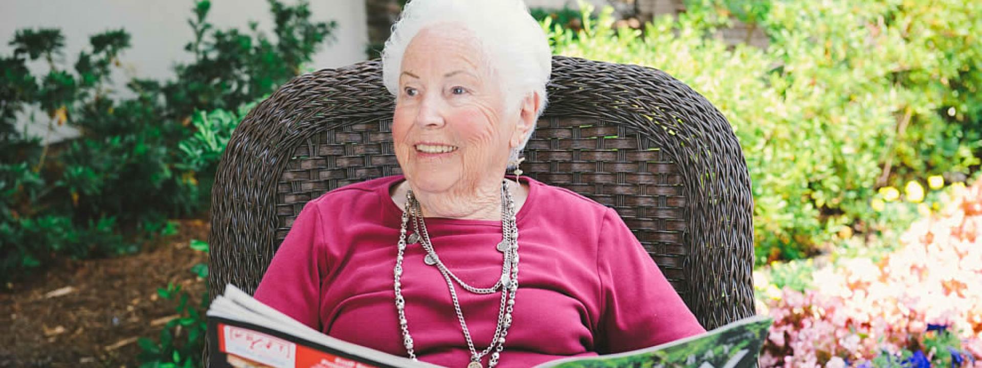 Smiling woman resident sitting in a brown rattan patio chair reading a magazine.