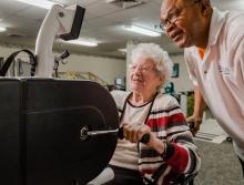 Woman resident on a stationary bike for therapy with staff standing next to her. 