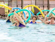 Six women in a pool doing aerobics holding foam pool noodles
