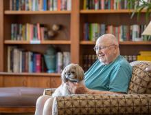 A smiling man sitting in the community library with his dog on his lap.