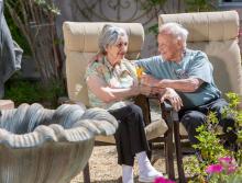 A smiling couple with their arms around each other, sitting outside next to a fountain and garden area. 
