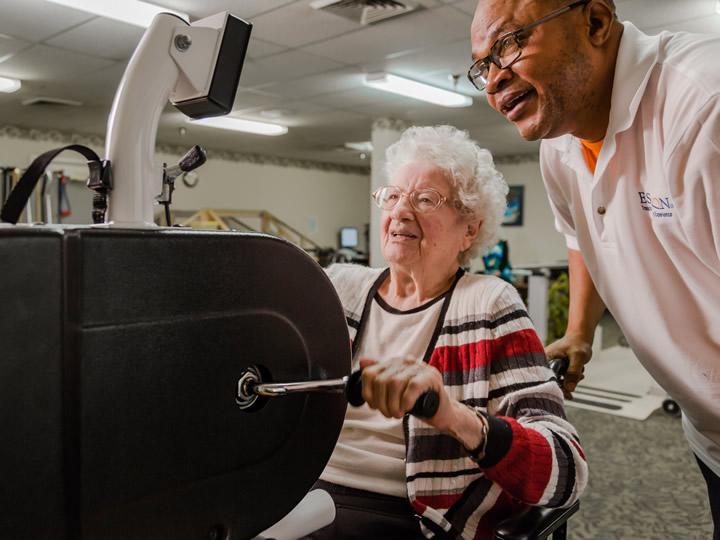Woman resident on a stationary bike for therapy with staff standing next to her. 