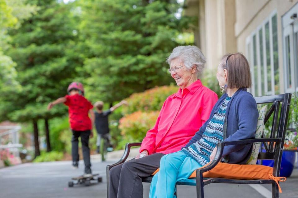 Two women resident sitting outside on a bench while two children are skating on their skateboards.