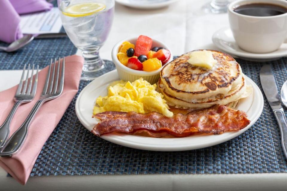 A breakfast plate with scrabbled eggs, bacon, pancakes and fresh fruit.