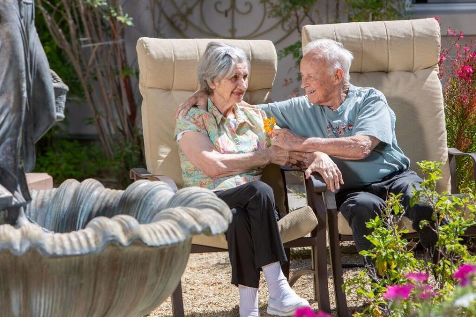 A smiling couple with their arms around each other, sitting outside next to a fountain and garden area. 