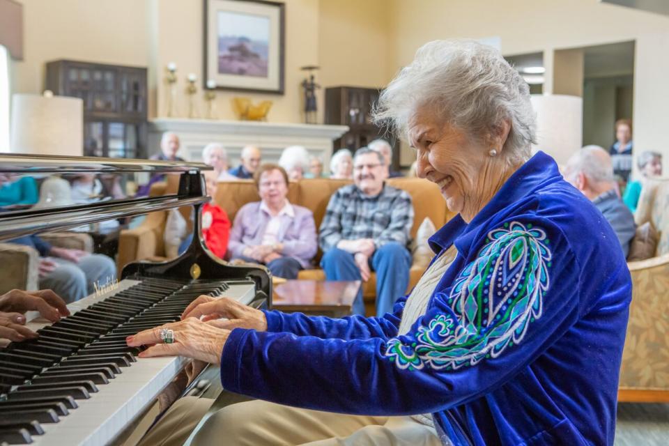 A smiling woman playing the piano for a group of residents.