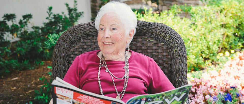 Smiling woman resident sitting in a brown rattan patio chair reading a magazine.