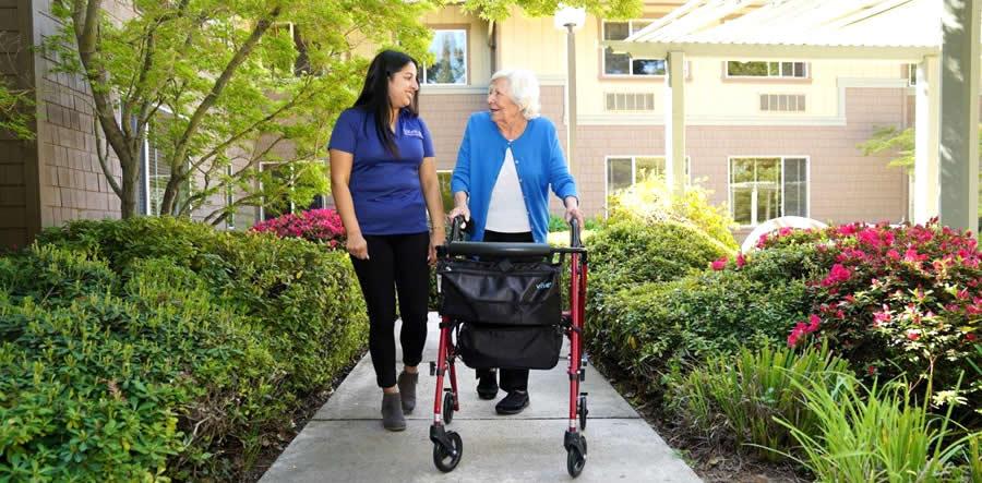 A caregiver walks alongside a woman using a walker.