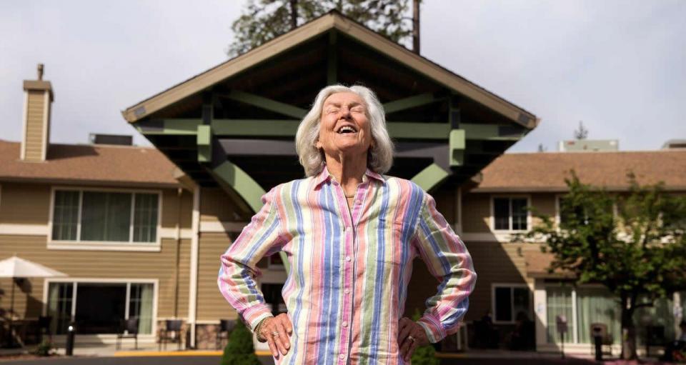 A woman standing outside enjoying the sun, with her hands on her hip, eyes closed with a big smile.