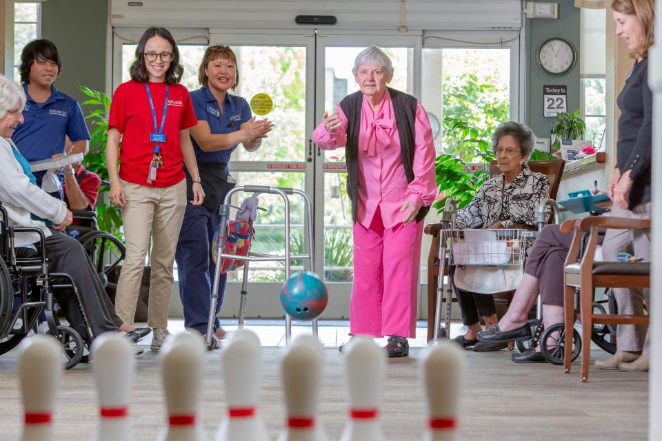 Resident woman throwing a bowling ball to knock down the bowling pins, with other residents and staff cheering her on.