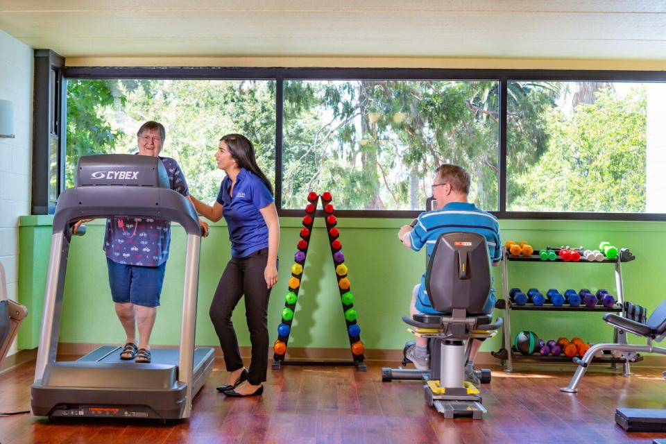 A woman on a treadmill and a man on a stationary bike in the exercise room with a staff member