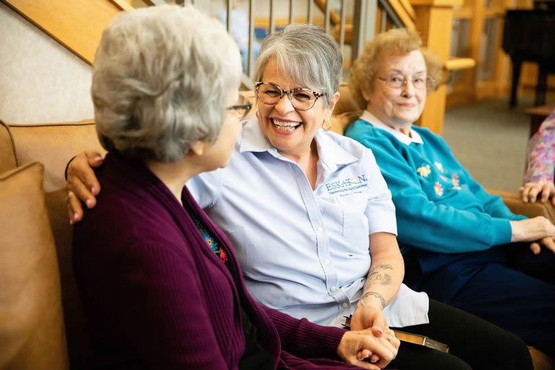 Eskaton Chaplin sitting on a couch smiling with two residents; holding one hand.